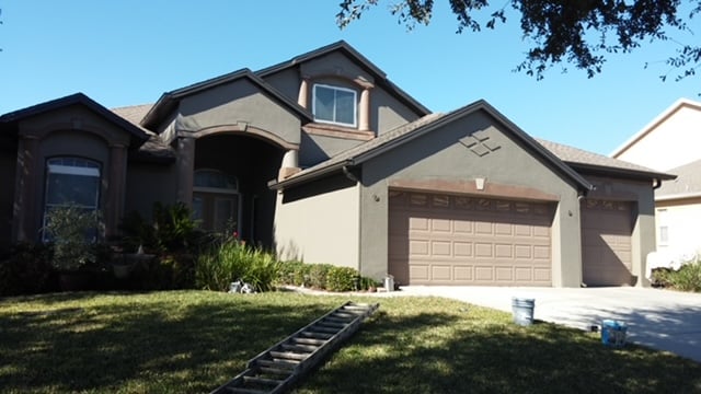 Gray stucco two-story home mid-repaint, with a ladder and paint buckets on the front lawn